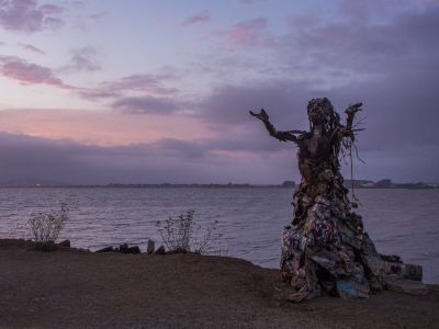 a sunset at albany bulb overlooking the san francisco bay with a statue of a person at the foreground. the person is made of found materials and appears to have arms outstretched with palms facing upwards. the person appears to be wearing a long flowing skirt